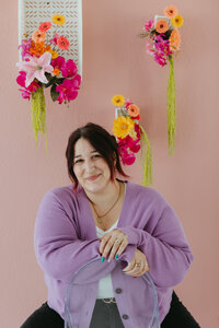 A close-up portrait of a person with dark purple hair in a loose updo, smiling warmly at the camera. They wear a lilac cardigan over a white top, with gold jewelry and colorful nail polish. The background features a pink floral wall with hanging blooms in shades of pink, orange, and yellow