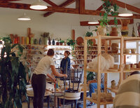 A 120 film photograph capturing the warm and inviting atmosphere of a pottery studio in San Diego during a branding session. The image features a duo engaging with students and working in the studio, surrounded by shelves of colorful handmade ceramics, clay supplies, and lush greenery. The natural light and wood accents highlight the creative and collaborative energy of the space, showcasing the artistry and charm of this pottery studio.