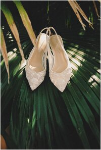 A bride's shoes sit on a palm branch in the shade