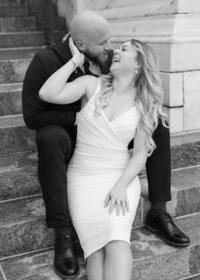 A couple sitting on the steps of Byron White Courthouse in Denver laugh at each other during their engagement portraits.