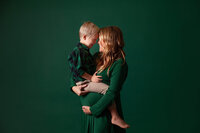 A Jacksonville maternity photographer captures a woman with long hair standing against a green background, holding a smiling young boy in her arms. Both are dressed in green clothing. The boy rests his forehead on hers with a joyful expression, and the woman looks at him lovingly.