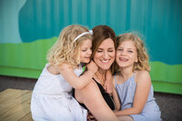 A woman sits smiling with her eyes closed, embraced by two young girls with curly blond hair. They wear pastel dresses against a colorful backdrop.