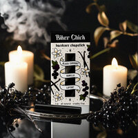 A box of Biker Chick Chapstick is illustrated with traditional tattoo art style icons of vanilla, black currents, and sits on a shiny black table with smoke, candles, and black currents sitting around it.