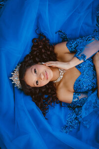 A young woman in a blue quinceañera dress lies on the ground with a tiara on her head during a portrait session at Mercer Botanic Garden in Humble, Texas.