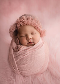A newborn baby sleeps in a pink swaddle and bonnet in a studio