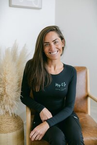 A woman with long brown hair wearing a black outfit sits smiling on a brown chair with her hands clasped, next to a decorative arrangement of light-colored pampas grass at a Maine medical aesthetics clinic.