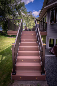 TimberTech Advanced PVC deck stairs with integrated riser lights and bronze aluminum picket railing with post cap lights on a sunny summer day. 