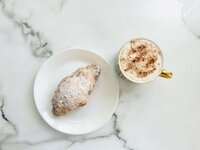 Image shows the top view of a cup of frothy coffee topped with a dusting of cocoa powder next to an almond croissant dusted with powdered sugar on a plain white saucer. Both are set against a grey and white marble countertop