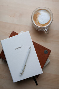 White pen and journal on top other journals next to a mug of Chai latte
