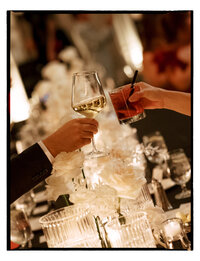 April M Payne Photography taken of two hands clinking glasses at a wedding reception, with a beautiful table setting of white flowers and soft lighting in the background.