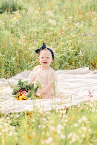A young toddler, wearing white and a blue bow, holds a bouquet of flowers while sitting on a white blanket in a field of flowers.