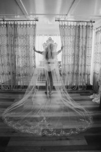 Black and white bridal boudoir photo of a bride in a long lace-edged cathedral veil, standing before patterned curtains with sunlight streaming in, taken as part of her intimate wedding morning portraits. Photo by Auburn Blue Photography.
