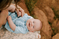 baby boy wearing  denim romper lays on top of moms thighs while smiling on a summer day
