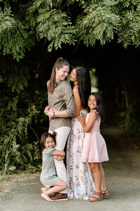 a young lesbian LGBTQ+ couple standing by an arbor, hugging one another while their two daughters hug ones leg and the others side