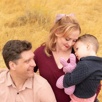 Mom and dad with 2 year old child and his stuffed animal. Dried yellow grasses in the background.
