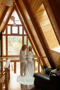 Bride standing inside a wooden cabin, framed by large windows, preparing for her destination elopement.