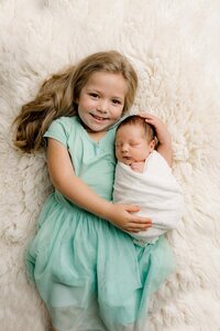 toddler sister dressed in teal poses on a cream fur holding her days old newborn brother during this sibling portion of their charlotte newborn session