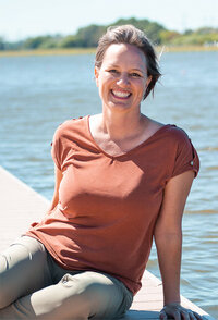 The photo shows a woman seated on a wooden dock, smiling at the camera with a relaxed, joyful expression. She is casually dressed in a burnt orange t-shirt with button details on the shoulder and light khaki pants. Her hair is styled in a short, tousled look. Behind her, the serene backdrop features a calm body of water reflecting the bright daylight and a clear blue sky with no visible clouds. The setting suggests a warm, pleasant day, likely at a lake or a calm river.
