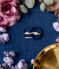 Wedding rings surrounded by florals on a blue backdrop.