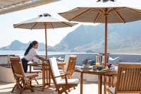 Waitress setting luxury dining table on a seaside terrace with parasols and ocean views.