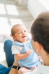 Austin Texas Portrait Photographer | A newborn baby boy dressed in a blue romper is held by his father. He faces him and smiles. 