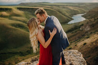 A man dressed in a nice suit holds the face of his girlfriend or fiance and kisses her while slightly dipping her. She is wearing a bright red dress and is holding his arm. Behind them is a valley with a river winding through it.