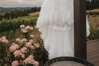 Elegant white wedding dress hanging beside blooming pink roses on a vineyard deck, with rolling hills of the Oregon wine country in the background, captured by a Portland wedding photographer specializing in Pacific Northwest vineyard weddings