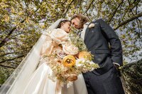 A close-up of a bride and groom touching foreheads beneath a flowering tree, with a large bouquet of peach, ivory, and lavender blooms in the foreground, captured by a UK wedding photographer.