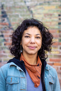 A headshot of Mexican-American artist Krissy Ramirez  with aged painted bricks in the background
