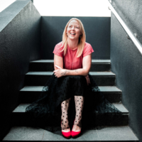 Image of Hayley Maxwell a copywriter in NZ sitting on steps smiling in a pink top and black skirt.
