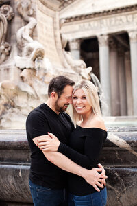 A couple wearing black tops with jeans embracing in front of the fountain at the Pantheon. Taken by Rome Photographer, Tricia Anne Photography