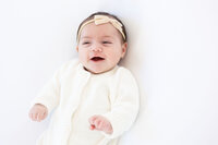 Young newborn laughing while wearing a white knit jumper and a matching bow.