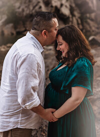Toddler stands at the end of a long twisting lane that curves behind her. There's snow on the ground and tall brown grass behind her with trees in the background. She is wearing a tan coat and white snow hat with a pompom and is smiling big at the camera in this family photoshoot.