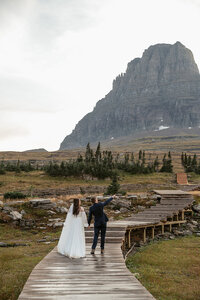 A bride and groom walk hand in hand along a wooden boardwalk beneath towering mountain peaks in Glacier National Park, captured by Sydney Breann Photography during their adventurous Montana elopement.