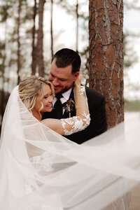 Bride and groom embracing beneath tall pine trees, her veil sweeping around them during an intimate wedding portrait session in Florida.