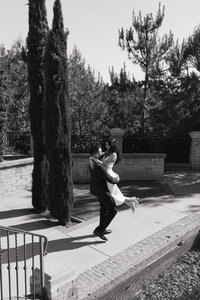 Groom lifts smiling bride into the air as they share a joyful moment outside, surrounded by trees and cypress.