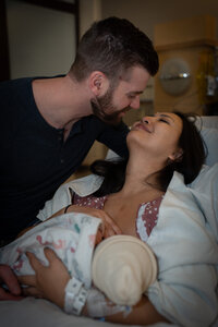 a husband is about to kiss his wife who is holding  their new baby in the hospital bed.