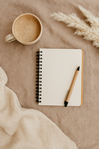 Flat lay photo with a cozy beige notebook, pen, warm coffee mug, and pampas grass arranged on a soft fabric background, representing a calming self-love journaling scene.