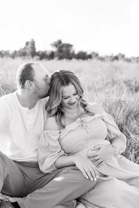 A man kissing his pregnant wife on the head while sitting in a field in Panama City Beach.