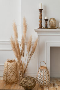A stylish interior scene featuring a white fireplace adorned with two wooden candlesticks and decorative items. Beside it are tall dried pampas grasses, woven baskets, and lanterns on a wooden floor. The aesthetic is natural and minimalist.