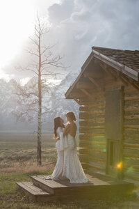 Brides hug and dance on  the porch of a rustic cabin  and the sun is setting behind the Grand Tetons behind them.