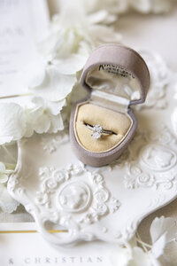 A diamond ring in a beige velvet box, resting on a textured white surface with a small cluster of white flowers beside it.