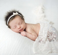 Newborn girl laying on belly on a white patterned blanket and covered in a white lace throw.
