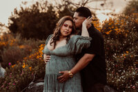 Expecting couple embracing in a wildflower field at sunset during a maternity photoshoot, with the woman in a sage green dress and the man holding her from behind