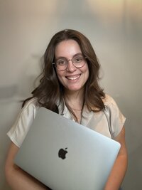 Woman with glasses and brown hair, smiling while holding a laptop, representing Molli Gorrell working as a virtual assistant and web designer.