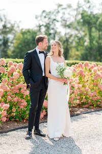 A bride peers over her shoulder as her groom stands behind her and they pose in front of pink hydrangeas.