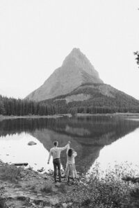 couple cheering for their engagement on the shore of lake sherburne in glacier national park