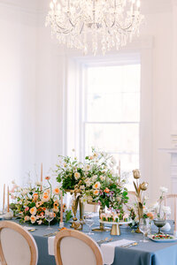 Elegant table setting with floral arrangements, blue tablecloth, chandelier above.