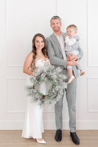 family of three mom holding a wreath and dad holding baby christmas card photo by miami christmas mini session photographer msp