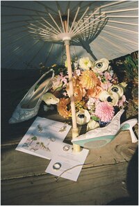 A bride's bouquet, shoes and rings lay on the boardwalk under an umbrella